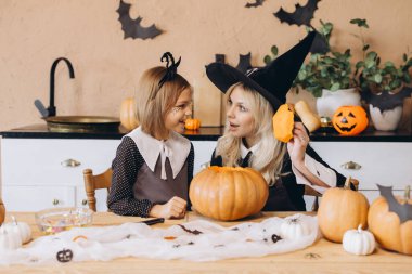 Mother wearing witch hat and daughter carving pumpkins together, preparing for Halloween party celebration at home