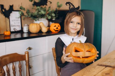 Little girl dressed in a witch costume, holding a carved pumpkin and celebrating Halloween joyfully at home with festive decorations