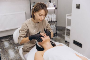 Professional beautician performing facial massage to client lying on massage table in modern beauty salon
