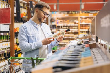 Smiling man choosing a bottle of white wine in a supermarket drinks section, reading the label and pushing a shopping cart