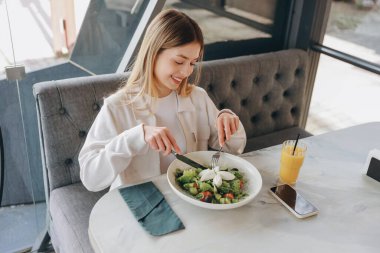 Happy young woman is about to eat a fresh salad in a modern restaurant, with a smartphone and a glass of orange juice on the table