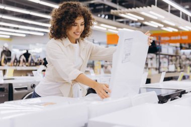 Smiling woman selecting a washing machine while examining specifications in a home appliance store, surrounded by various options