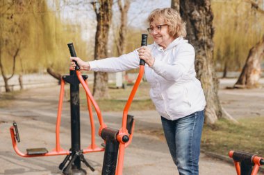 Elderly woman using elliptical cross trainer in outdoor park gym, enjoying healthy lifestyle and active aging