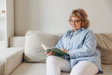 Elderly woman wearing eyeglasses enjoying a book while relaxing on a comfortable couch in her living room