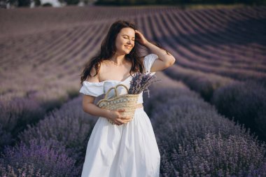 Young woman holding a straw bag and a bouquet of lavender in a lavender field, enjoying the scent and the beautiful landscape