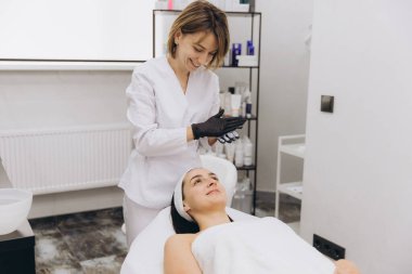 Smiling beautician wearing black gloves preparing charcoal face mask for her client lying on massage table in beauty salon
