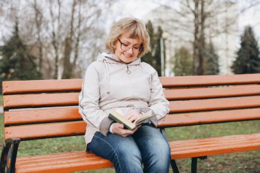 Elderly woman sitting on a park bench, happily reading a book while enjoying a peaceful moment in nature during her leisure time outdoors