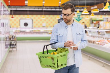 Customer writing on notepad while carrying a shopping basket full of fresh vegetables inside a supermarket