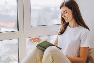 Smiling woman sitting by the window, wearing casual clothes and reading a book in natural light