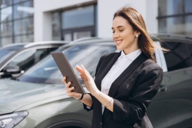 Saleswoman using digital tablet and working at car dealership, checking online catalog or inventory