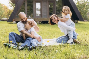 Smiling parents and children relaxing on grass in front of their a frame house, enjoying quality time together