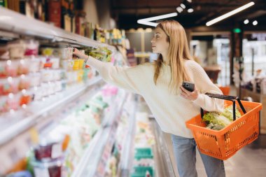 Dondurulmuş raflardan süt ürünlerini seçen genç bir kadın, elinde alışveriş sepeti ve akıllı bir telefonla, parlak ışıklı bir süpermarkette