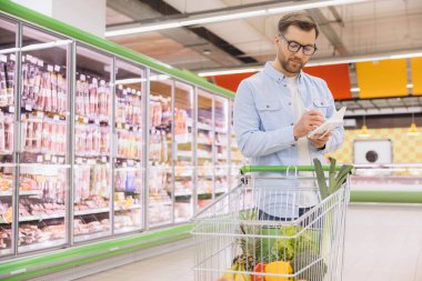 Man writing notes on a notepad while pushing a shopping cart filled with groceries through the bustling supermarket aisles