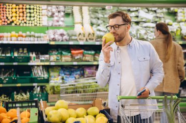 Customer smelling a green apple while holding a shopping list and pushing a shopping cart in a supermarket