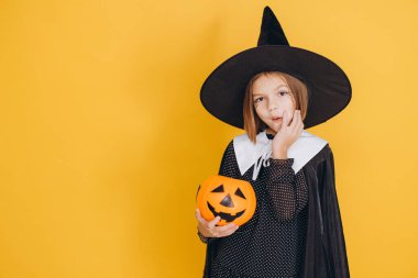 Cute little girl wearing witch costume holding pumpkin basket posing on yellow background for Halloween party