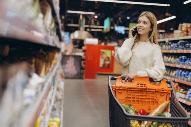 Alışveriş arabasını iten ve marketten alışveriş yaparken akıllı telefondan konuşan genç bir kadın.