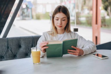 Concentrated young woman enjoying a book and drinking orange juice while relaxing in a modern cafe