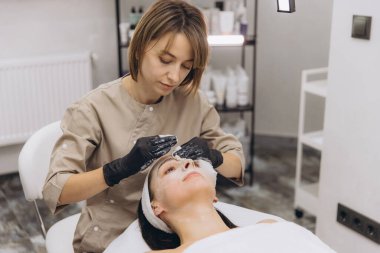 Professional beautician applying a rejuvenating facial treatment to a client relaxing on a clean bed in a modern beauty salon