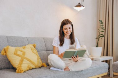 Young woman sitting on a sofa with crossed legs, smiling and reading a book, enjoying a quiet moment at home