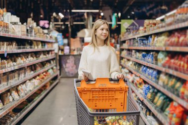 Elinde alışveriş listesi tutarken ve süpermarket koridorlarında yürürken alışveriş arabasını iten genç bir kadın.