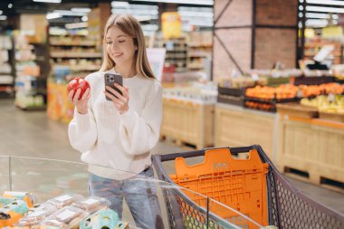 Akıllı telefon kullanan ve süpermarkette nar meyvesi seçen genç bir kadın, alışveriş konsepti.
