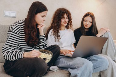 Three teenage girls are sitting on a sofa, using a laptop and eating popcorn, enjoying their time together
