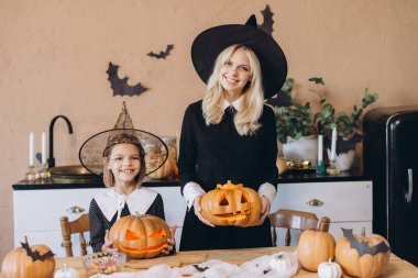 Mother and daughter smiling and wearing witch costumes holding carved pumpkins in a decorated kitchen for Halloween