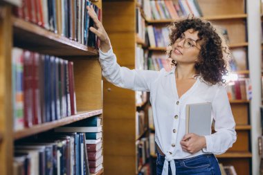 Young woman wearing eyeglasses selecting book from bookshelf in library, holding another book in her hand