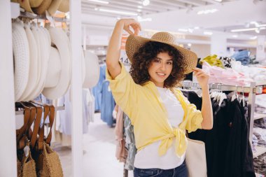 Smiling woman with curly hair trying on a stylish straw hat while enjoying a shopping experience in a vibrant fashion store