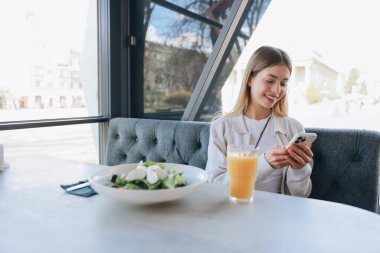 Smiling woman sitting in a modern cafe, enjoying a fresh salad and orange juice while using her smartphone for texting or browsing