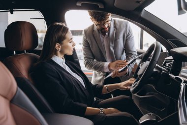Businesswoman testing new car features while sitting in driver's seat and listening to car salesman's explanations in a car dealership