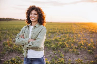 Confident young female agronomist standing with crossed arms in a soybean field at sunset, smiling