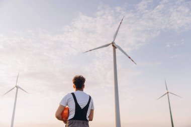 Arab engineer observing wind turbines in a wind farm during sunset, highlighting the concept of sustainable energy and innovation