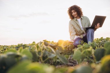 Young woman agronomist using a laptop while inspecting soybean crops in a vibrant field during a picturesque sunset