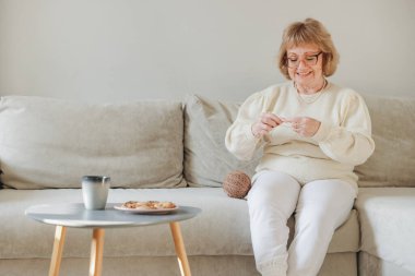 Elderly woman enjoying her retirement knitting on the sofa with a cup of coffee and cookies on the side table
