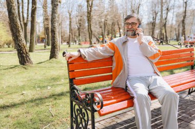 Bearded senior man wearing tracksuit is sitting on bench in park and talking on smartphone on sunny day