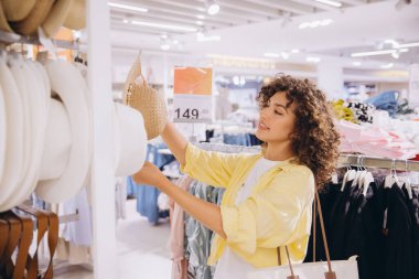 Smiling woman with curly hair browsing through hats in a vibrant clothing store, enjoying the shopping experience and exploring fashionable options