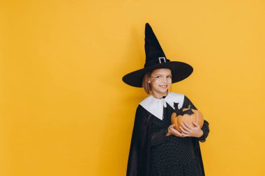 Young girl wearing Halloween witch costume holding pumpkin with bat sticker posing on yellow background
