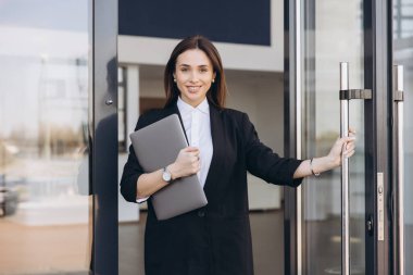 Smiling saleswoman holding a laptop is opening the door of a car dealership, ready to welcome customers