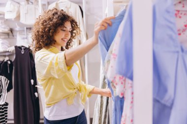 Smiling curly haired woman shopping for clothes, examining garments on a rack in a fashion store