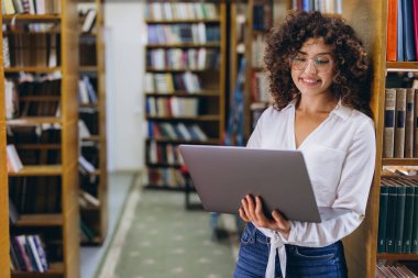 Young curly haired student wearing eyeglasses holding laptop smiling in university library between bookshelves