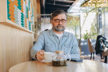 Bearded senior man enjoying a cup of tea in a bright, modern cafe, embodying a peaceful moment of relaxation