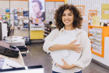 Smiling woman holding a smart home appliance in an electronics store, showcasing new technology and consumer choice