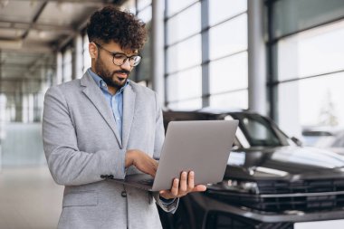 Car salesman working on laptop in front of new car, managing inventory and sales in modern dealership