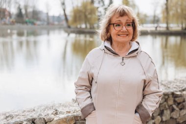 Elderly woman smiling and enjoying a peaceful walk along a serene park path, surrounded by the beauty of a tranquil lake and vibrant trees