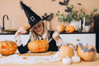 Smiling woman wearing a witch hat, joyfully carving pumpkins to create festive decorations for an exciting Halloween party