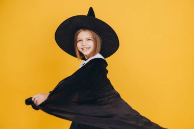 Happy child with witch hat and black cape having fun during Halloween celebration, waving her cloak on vibrant yellow background
