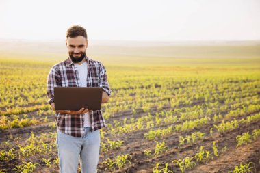 Bearded agronomist smiling while working on a laptop in a corn field during a stunning sunset, enjoying the beauty of rural life