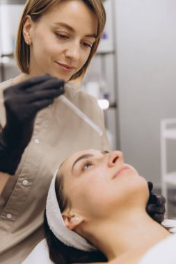 Beautician wearing black gloves applying serum on woman's face with a pipette, in beauty salon