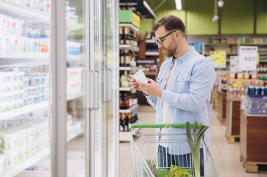 Man choosing groceries, reading product information on a package in front of refrigerated section in a supermarket